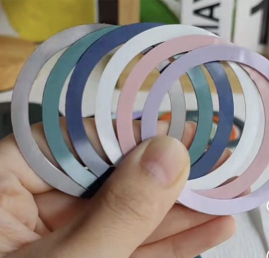 A hand holding a fan of colorful aluminum-clad magnetic ring plates with CD-texture finishes, anodized in various shades like blue, pink, silver, and purple.