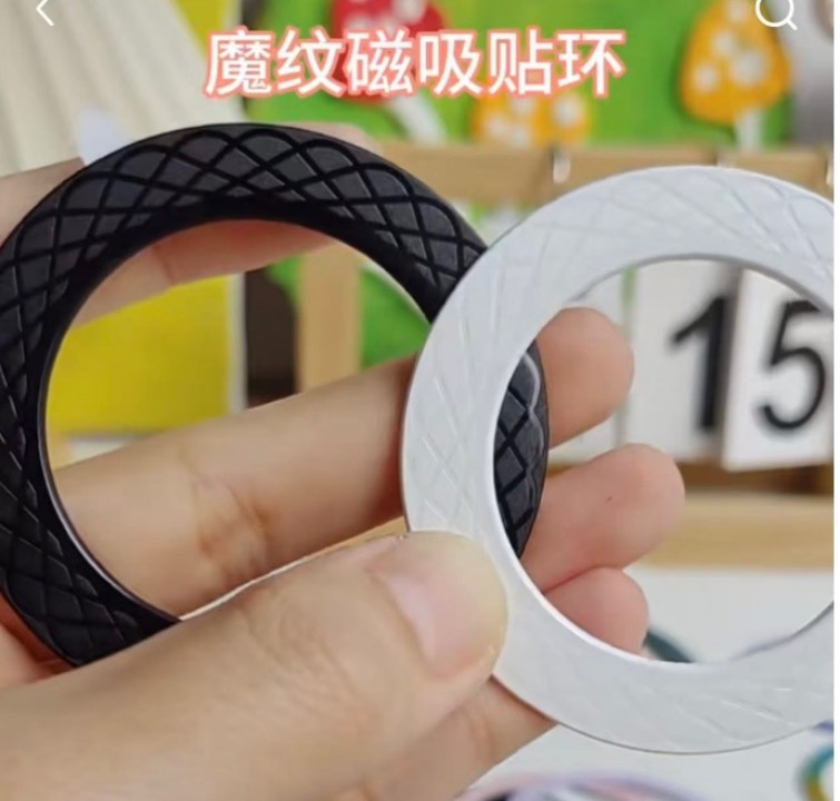 Hand holding two magnetic ring stickers in black and white, both with rhombus-embossed magic texture surfaces creating optical 3D effects.